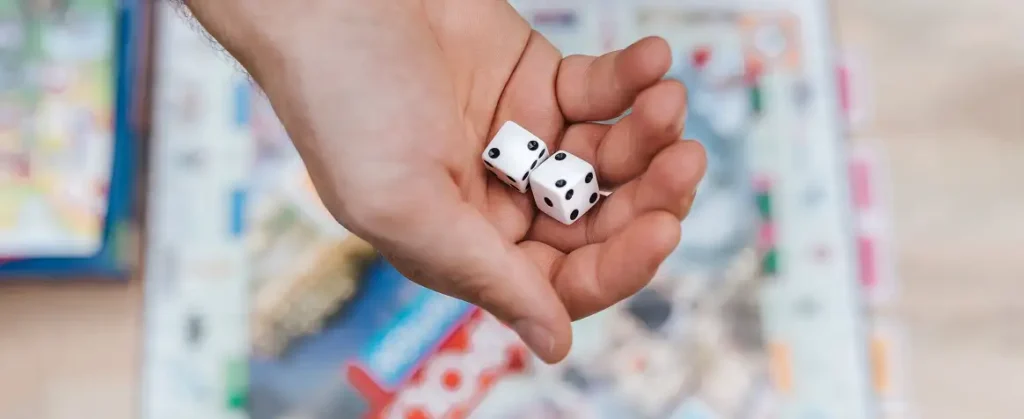 Zbliżenie na męską dłoń z dwiema białymi kostkami do gry na tle rozmytego pola gry Monopoly